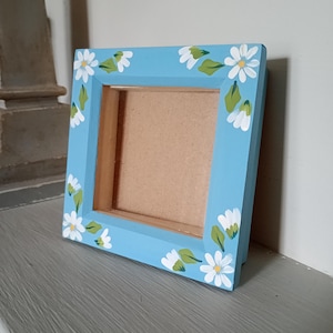 May include: A square, light blue picture frame decorated with hand-painted white daisies and green leaves. The frame has a wooden backing. The frame is designed to hold a photograph or artwork. The frame is on a grey surface.