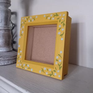 May include: A yellow picture frame with a white mount and a floral design painted around the edge. The flowers are white with green stems.