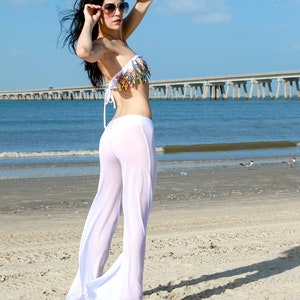 May include: A white strapless bikini top featuring a decorative fringe of multi-colored, pointed accents paired with sheer, floor-length white wide-leg trousers. The ensemble is worn with large, dark-tinted sunglasses with gold-toned frames.
