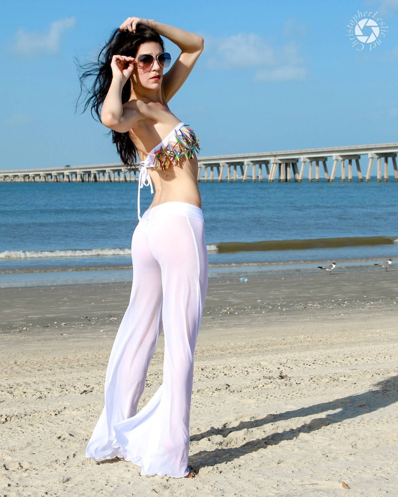 May include: A white strapless bikini top featuring a decorative fringe of multi-colored, pointed accents paired with sheer, floor-length white wide-leg trousers. The ensemble is worn with large, dark-tinted sunglasses with gold-toned frames.
