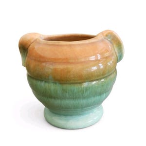 Puede incluir: Un jarrón de cerámica de dos tonos. La parte superior es naranja, que se mezcla con una sección media verde, con una base azul claro. El jarrón presenta dos asas curvas y una forma redondeada, adecuado para la decoración del hogar. Aproximadamente 15 cm de altura.