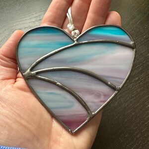 May include: Stained glass heart suncatcher with a silver-colored metal frame. The heart features a blend of blue, purple, and white glass, creating a soft, watercolor effect. A white cord is attached for hanging. A decorative item.