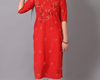 Puede incluir: Una kurta roja de manga larga con bordados dorados y negros. La kurta tiene un escote redondo y está decorada con motivos florales. La modelo lleva pantalones blancos y joyas de plata.