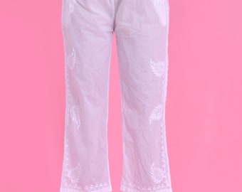 Può includere: Pantaloni bianchi ricamati con una vestibilità rilassata. I pantaloni presentano ricami floreali sui lati e sull'orlo inferiore. La modella indossa i pantaloni con sandali. Lo sfondo è rosa.