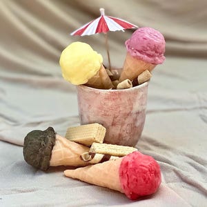May include: A terracotta pot filled with ice cream cones, some with yellow and pink scoops, and a miniature red and white striped parasol. Several cones with chocolate and red scoops are scattered around, with wafer biscuits.