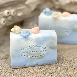 Può includere: Due saponette fatte a mano con un design a spirale blu e bianco. Le saponette sono decorate con conchiglie e il testo "Handmade Soap" è stampato sul davanti.