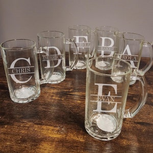 May include: Six clear glass beer mugs with handles. Each mug has a different letter etched on the front with a name underneath. The names are Chris, Derrick, Farrell, Baker, Amrola, and Evan.