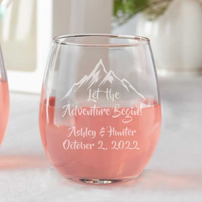 May include: A clear glass wine glass with a mountain design and the text "Let the Adventure Begin! Ashley & Hunter October 2, 2022" etched on the side. The glass is filled with a pink beverage.
