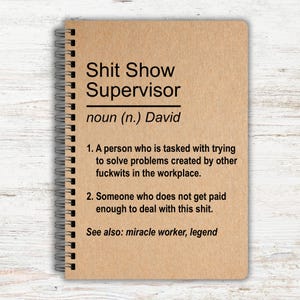 May include: A spiral-bound notebook with a brown cover. The text on the cover reads "Shit Show Supervisor" with a definition below. The definition includes the name "David" and two points about the role. The notebook is on a light wood-grain surface.