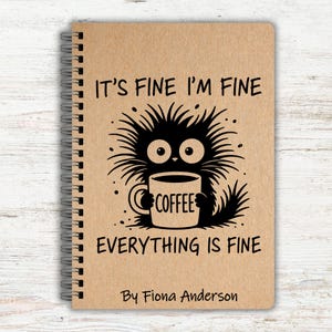 May include: A spiral-bound notebook with a brown cover. It features a black cartoon creature holding a coffee mug with the word "COFFEE". The text reads "IT'S FINE I'M FINE EVERYTHING IS FINE" and "By Fiona Anderson".