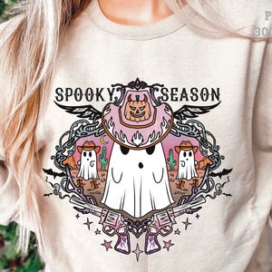 May include: Beige sweatshirt with a colorful "Spooky Season" graphic. The design features a ghost wearing a cowboy hat with a pumpkin, cacti, and revolvers. The graphic is surrounded by decorative elements.