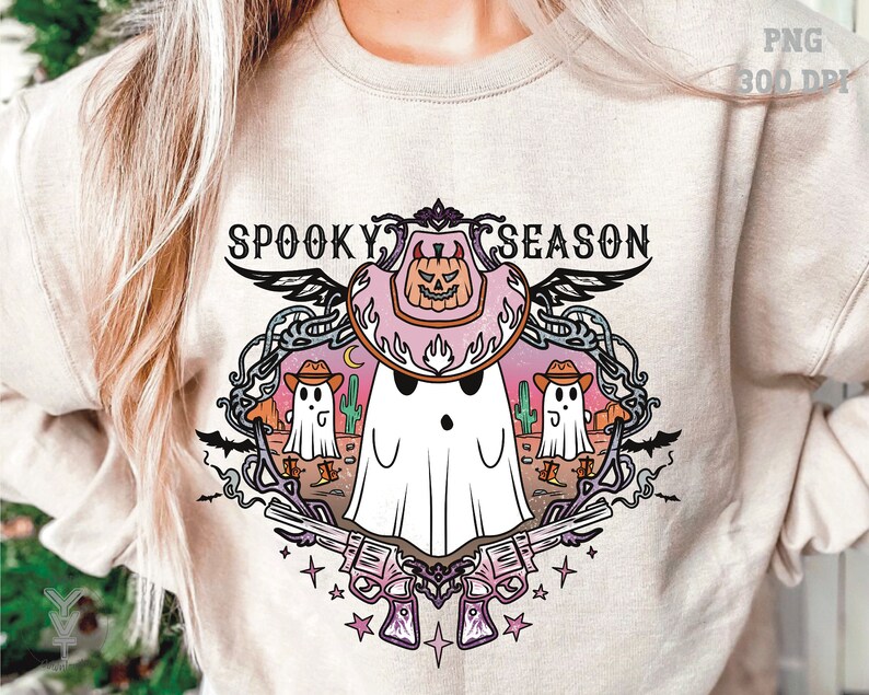 May include: Beige sweatshirt with a colorful "Spooky Season" graphic. The design features a ghost wearing a cowboy hat with a pumpkin, cacti, and revolvers. The graphic is surrounded by decorative elements.