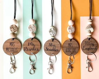 May include: Five wooden keychains with different teacher names engraved on them. Each keychain has a black lanyard with beads and a charm. The charms include a mushroom, a flower, and a leaf.