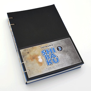 May include: A black leather-bound notebook with a blue and white cover. The cover features the text "SHIBAKU 3 ABHEBEN & FLIEGEN".