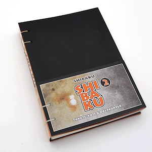 May include: A black leather-bound notebook with a title page featuring the text "SHIBAKU" in orange letters, a number "2" in a circle, and the text "TAKATE-KOTE & OBERKÖRPER" in black letters.