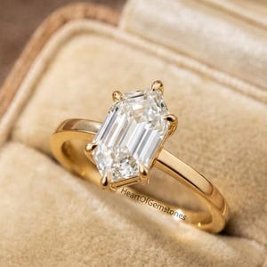 Puede incluir: Un anillo de compromiso de oro con una gran piedra preciosa clara con talla esmeralda. El anillo tiene una banda sencilla y se presenta en un joyero vintage forrado de terciopelo. El texto "HeartOfGemstones" es visible en el anillo.