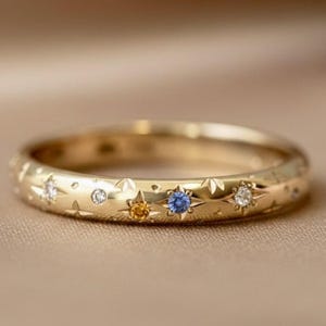 May include: A gold ring with a celestial design. The ring features small star and floral engravings, and is adorned with a variety of gemstones, including a blue sapphire, a yellow sapphire, and several diamonds. The ring is set against a neutral background.