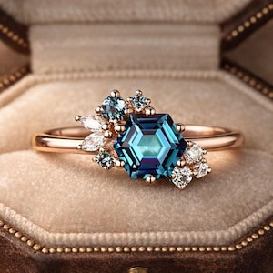 May include: A rose gold ring featuring a large, hexagonal teal gemstone as the centerpiece, surrounded by smaller blue and white accent stones. The ring is displayed in a brown velvet jewelry box. The text "HeartOfGemstones" is visible on the box.