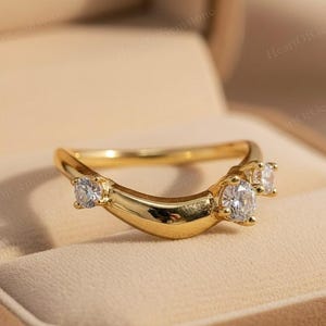 Puede incluir: Un anillo dorado con un diseño curvo, con tres piedras preciosas redondas y transparentes. El anillo se presenta sobre un fondo suave y neutro, destacando sus características elegantes y delicadas. El diseño del anillo es simple pero sofisticado.
