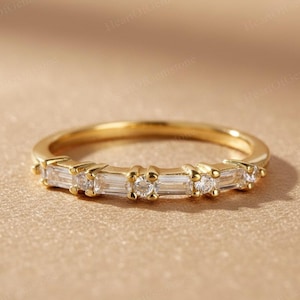 May include: A gold band ring featuring a series of alternating baguette and round-cut clear gemstones. The ring is set on a neutral-toned surface, highlighting the sparkle of the stones. A delicate piece of jewelry.
