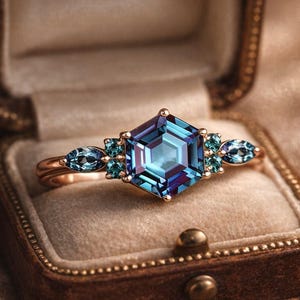 Op de afbeelding: Een gouden ring met een grote, zeshoekige, blauwgroene en paarse edelsteen als middelpunt. Kleinere blauwgroene edelstenen flankeren de centrale steen. De ring wordt gepresenteerd in een bruine sieradendoos met een beige interieur.