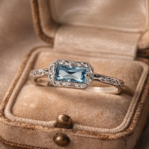 Könnte beinhalten: Ein silberner Ring mit einem rechteckigen, hellblauen Edelstein, umgeben von kleineren, klaren Steinen. Der Ring wird in einer Samt-Ringbox präsentiert. Das Band hat ein verziertes, graviertes Design. Ein klassisches Schmuckstück.