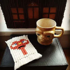 May include: A white crocheted coaster with a red, yellow, and blue geometric design sits on a black leather surface next to a beige ceramic mug with a face sculpted on the front.