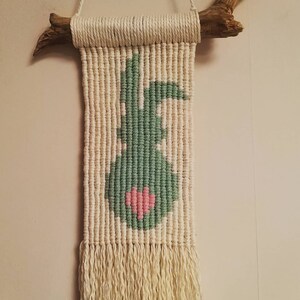May include: A macrame wall hanging with a green bunny silhouette and a pink heart. The hanging is attached to a natural wood branch.