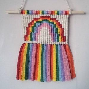 May include: A rainbow-colored macrame wall hanging with a white background and fringe. The rainbow is made of different colored yarn and is in the shape of an arch.