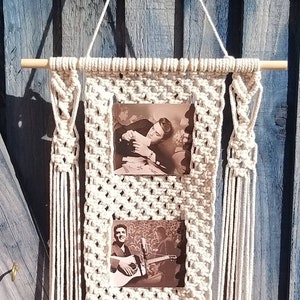 May include: A macrame wall hanging with two photo slots. The wall hanging is made of natural cotton cord and has a wooden dowel at the top and bottom. The photos are of Elvis Presley.