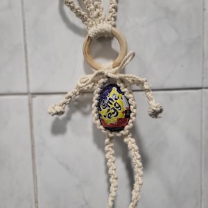 May include: A white crocheted bunny with a wooden ring and a Cadbury Creme Egg inside. The bunny is hanging from a white wall.