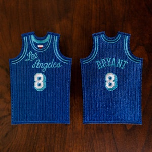 Puede incluir: Dos camisetas de baloncesto azules con costuras blancas y turquesa. La camiseta delantera tiene "Los Angeles" y el número "8" en la parte delantera. La camiseta trasera tiene "BRYANT" y el número "8" en la parte trasera.