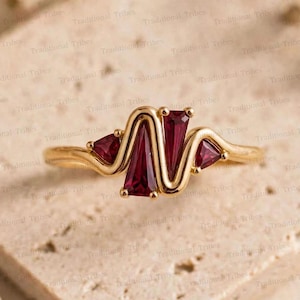 May include: A gold ring featuring a zigzag band design set with four deep red, trapezoidal-cut gemstones. The metal band follows a continuous wave pattern, securely holding the faceted red stones in a symmetrical arrangement across the top.