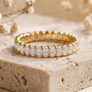 Könnte beinhalten: Ein goldener Eternity-Ring mit einer durchgehenden Reihe von oval geschliffenen, klaren Steinen in einer gemeinsamen Krappenfassung. Der Ring liegt auf einem strukturierten, hellen Steinblock und zeigt eine gleichmäßige Anordnung der Edelsteine.