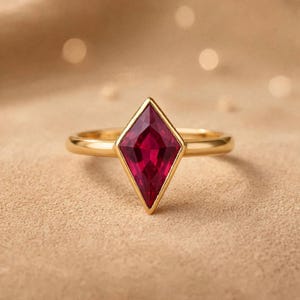May include: A gold ring featuring a kite-shaped, deep red gemstone set in a smooth metal band. The faceted stone is positioned vertically, showcasing a vibrant crimson hue and a polished gold setting that encircles the gem's sharp edges.