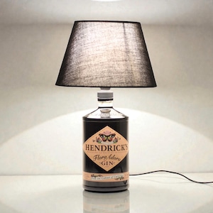 Può includere: Una lampada da tavolo unica realizzata con una bottiglia di gin Hendrick's riciclata. La bottiglia nera presenta un'etichetta con il nome del marchio e un'illustrazione di farfalla. È sormontata da un paralume nero e da un portalampada argentato.