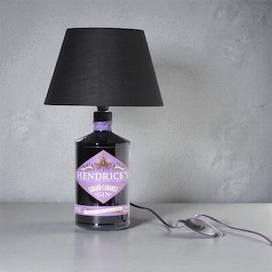 Può includere: Lampada da tavolo con paralume nero, realizzata con una bottiglia di gin. La bottiglia è scura con un'etichetta viola che recita "Hendrick's Grand Cabaret Gin". Un cavo nero con interruttore è collegato. Arredamento per la casa.