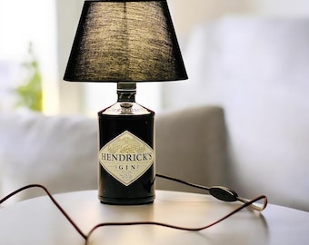 May include: A black lampshade with a black cord sits atop a clear glass bottle with a black label that reads "Hendrick's Gin".