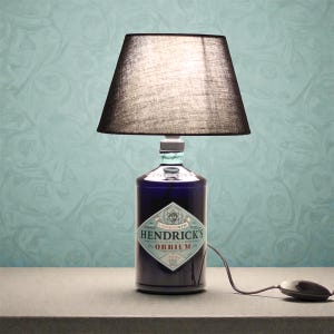 Può includere: Una lampada da tavolo unica realizzata con una bottiglia di gin Hendrick's Orbium riciclata. La bottiglia blu scuro presenta un'etichetta con il nome del marchio. La lampada ha un paralume nero e un cavo di alimentazione con interruttore.