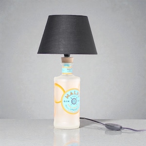 Può includere: Una lampada da tavolo realizzata con una bottiglia di gin riciclata. La bottiglia ha un'etichetta con il testo "MALFI" e "GIN". La lampada ha un paralume nero e un tappo di sughero. La lampada è collegata a un cavo di alimentazione.