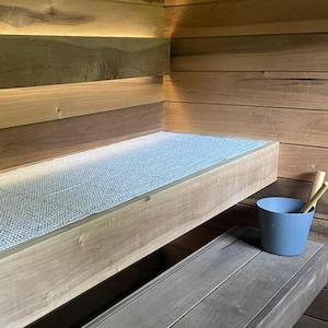 May include: Interior view of a sauna with wooden benches and walls. A light blue mat covers the top bench. A small blue bucket with wooden ladles sits on a lower bench. The wood has a natural, light brown color.