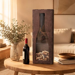 May include: A tall, rectangular, dark brown wooden box with a clear front panel shaped like a wine bottle, containing a collection of natural corks. The box stands upright on a wooden surface next to a bottle of red wine and a glass vase with greenery.