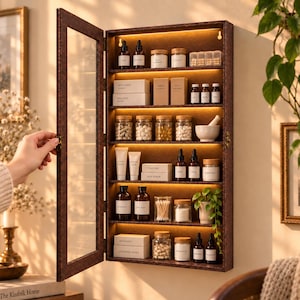 May include: A wall-mounted medicine cabinet with a dark brown textured frame and a glass door. The interior features five shelves illuminated by integrated LED lighting, holding various glass bottles, jars, boxes, and a small potted plant.