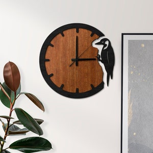 Puede incluir: Un reloj de madera marrón y negro con una silueta de pájaro carpintero negro. El reloj está colgado en una pared blanca con una cómoda de madera marrón y una planta verde en primer plano.