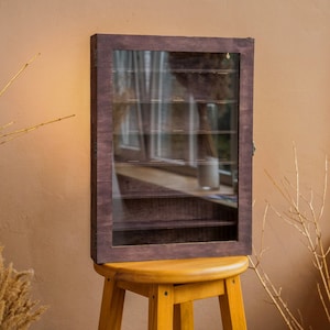 Puede incluir: Una vitrina de madera marrón oscuro con estantes de vidrio. La vitrina tiene un diseño sencillo y es perfecta para exhibir objetos pequeños.