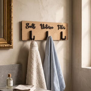 May include: A rectangular light wood wall-mounted rack featuring three black metal hooks and three black script names, Scott, Victoria, and Piter, mounted above each hook. A textured cream towel and a blue towel hang from the hooks.