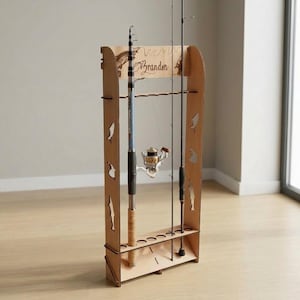 May include: A light-colored wooden fishing rod rack featuring cutouts of fish shapes on the side panels. The top horizontal panel is engraved with the name Brandon and a fish illustration. The rack holds two fishing rods with reels.