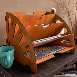 May include: A wooden magazine rack with a decorative cut-out design. The rack has three shelves and is made of light brown wood. It is sitting on a dark brown table.