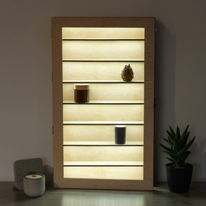 Puede incluir: Una vitrina de madera con estantes de vidrio y luces LED. La vitrina tiene seis estantes y está iluminada desde atrás. Hay un pequeño frasco en un estante y un pequeño objeto decorativo en otro estante.