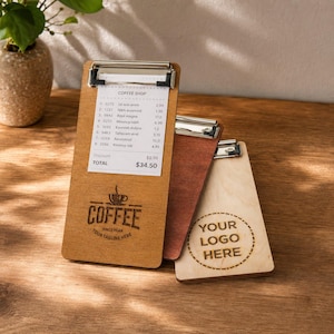 Op de afbeelding: Drie rechthoekige houten klemborden met metalen clips. Het voorste bord is lichtbruin met een koffiebon en een koffiekopje. Het middelste is donkerbruin en het rechterbord is licht hout met de tekst YOUR LOGO HERE erop gegraveerd.